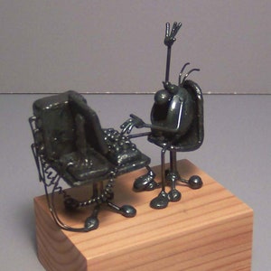 May include: A metal sculpture of a person sitting at a computer desk, with one hand raised in the air. The sculpture is painted black and is sitting on a wooden base.