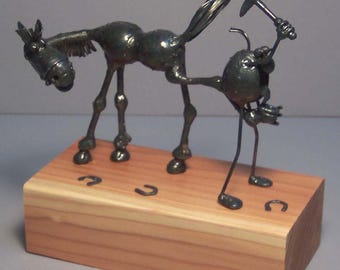 May include: A whimsical metal sculpture of a horse being shoed by a farrier. The dark metal figures stand on a rectangular wooden base with horseshoe imprints. The horse is bent over, and the farrier is hammering a horseshoe.
