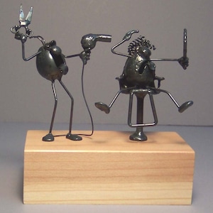 May include: A metal sculpture of a hairstylist cutting the hair of a customer. The hairstylist is holding scissors and a hairdryer, while the customer is sitting in a chair and looking in a mirror. The sculpture is on a wooden base.