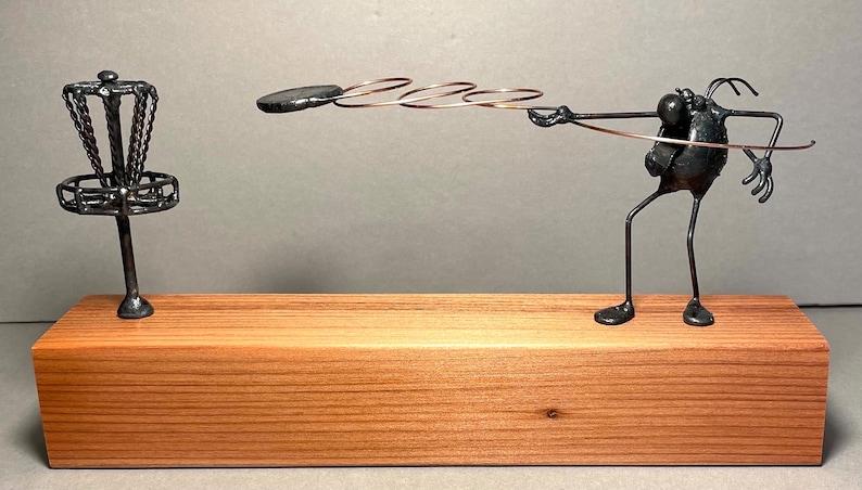 May include: A whimsical metal sculpture depicting a disc golf scene. A stylized figure made of dark metal wire throws a disc towards a miniature disc golf basket, also crafted from dark metal. The sculpture is mounted on a light brown wooden base. The artwork showcases intricate details and a playful design, suitable for disc golf enthusiasts or as a unique decorative piece.