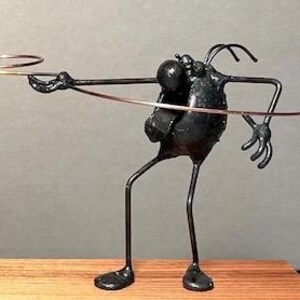 May include: A black metal sculpture of a stylized figure with long limbs and a round head. The figure is holding a ring in its outstretched hand.