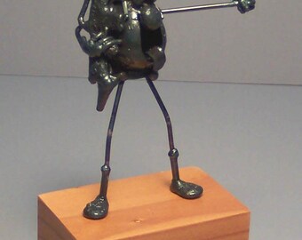 May include: A quirky metal sculpture of a figure with a rifle and pointing arm. The dark metal figure stands on thin legs atop a small, rectangular wooden block. The figure's form is abstract and stylized.