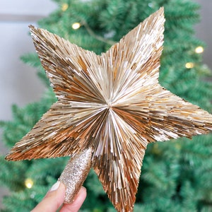 May include: A gold star tree topper with a sparkly finish. The star is made of thin, metallic strands that create a fluffy, textured look.