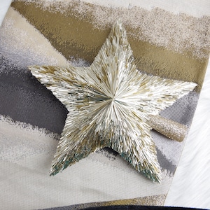 May include: A decorative star made of metallic gold rods, resting on a textured runner with gold, gray, and white brushstroke patterns. A smaller, glittery gold accent is attached to the star.