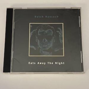 Può includere: Un compact disc in una custodia di plastica trasparente con copertina nera. La copertina presenta il testo Butch Hancock in alto e Eats Away The Night in basso. Una cornice quadrata centrale contiene un'illustrazione monocromatica di una persona con occhiali da sole e mani sollevate vicino alla testa.