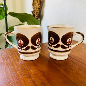 May include: Two white ceramic mugs with brown and red floral designs. The mugs have a brown rim and a brown base with speckles.