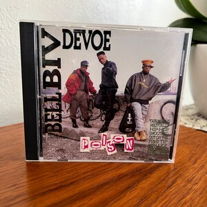 Könnte beinhalten: Ein CD-Gehäuse mit schwarzem Rücken und einer transparenten Vorderseite. Die Vorderseite zeigt ein Foto von drei Männern, die vor einer mit Graffiti besprühten Wand stehen. Der Albumtitel "Poison" ist in pinken und weißen Buchstaben gedruckt. Die Albumkünstler "Bell Biv DeVoe" sind in schwarzen Buchstaben gedruckt. Der Text "Our music is mentally hip-hop smoothed out on the R&B tip with a pop edge" ist auf der rechten Seite des Covers gedruckt.