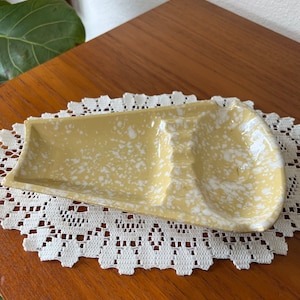 May include: A yellow ceramic ashtray with white speckles. The ashtray has three indentations for cigarettes.