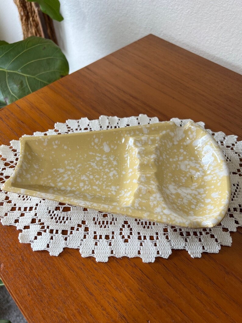 May include: A yellow ceramic ashtray with white speckles. The ashtray has three indentations for cigarettes.