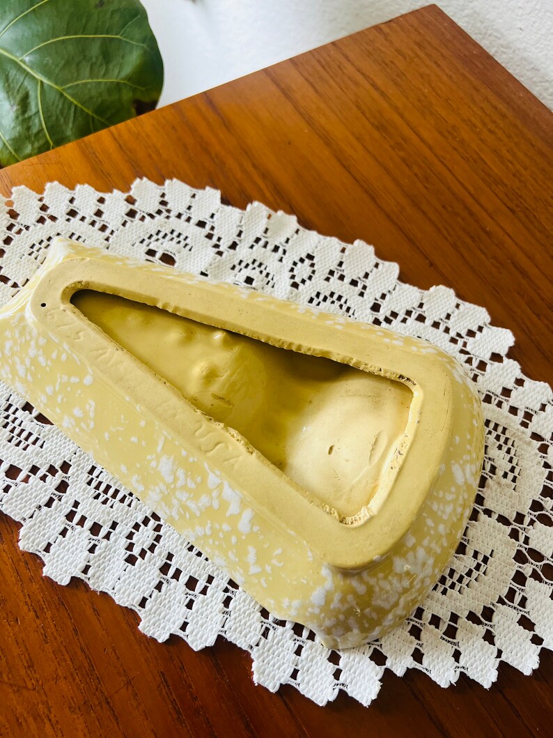 May include: A yellow ceramic ashtray with white speckles. The ashtray is shaped like a triangle with a rounded bottom. The inside of the ashtray is empty.