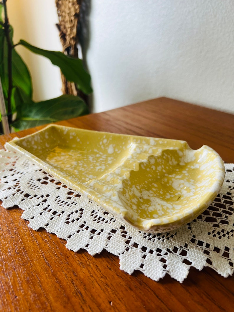 May include: A yellow ceramic ashtray with white speckles. The ashtray has two compartments for cigarettes.