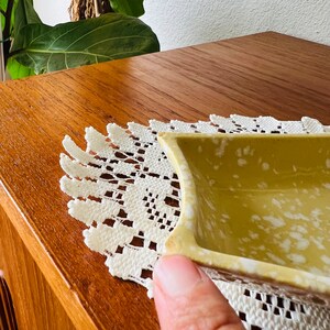 May include: A small, square, yellow ceramic dish with white speckles. The dish is sitting on a white crocheted doily.