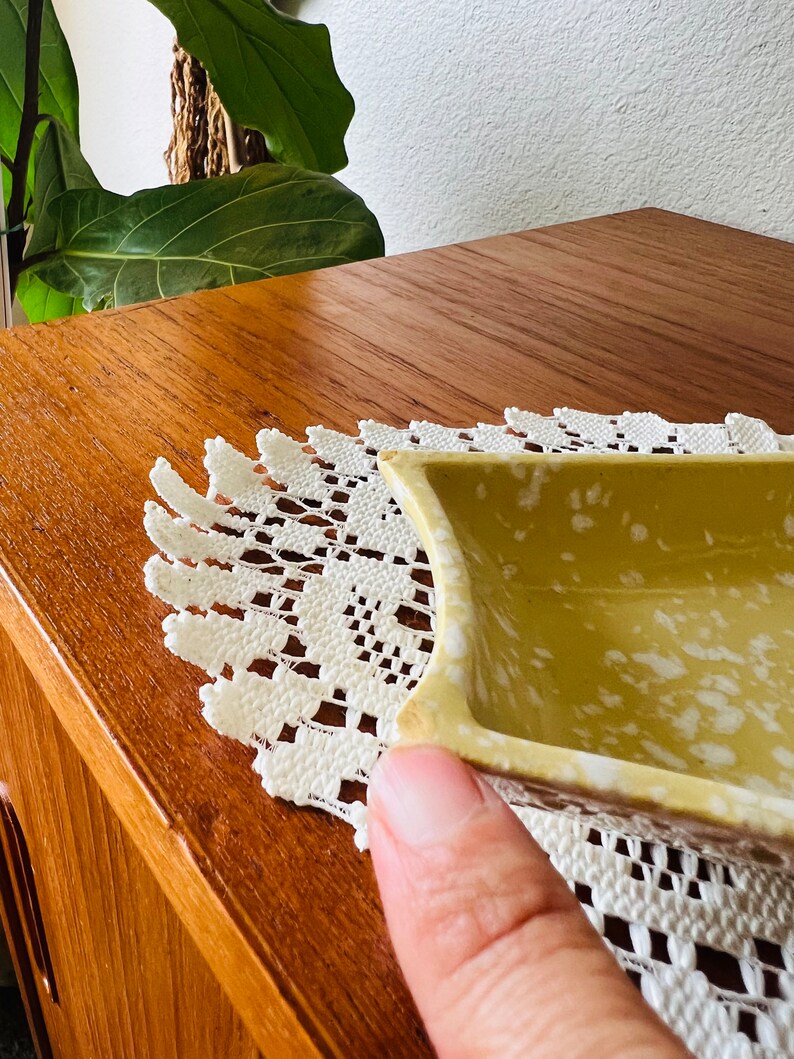 May include: A small, square, yellow ceramic dish with white speckles. The dish is sitting on a white crocheted doily.