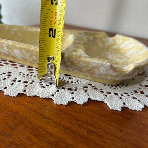 May include: A yellow and white ceramic ashtray with a divided section. The ashtray is 2 3/8 inches wide.