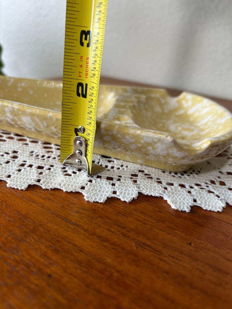 May include: A yellow and white ceramic ashtray with a divided section. The ashtray is 2 3/8 inches wide.