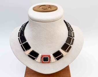 Puede incluir: Un collar de madera negro y blanco con un colgante cuadrado rojo y negro. El collar está hecho de cuentas de madera rectangulares con un alambre plateado que las atraviesa.