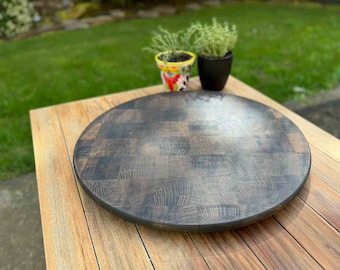 May include: A round, dark-stained wooden cutting board with a butcher block design. The board is placed on a wooden table. Two small potted plants are in the background. The board is approximately 40 cm in diameter.