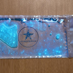 May include: A clear plastic package with a blue, glowing interior. The package contains a product with a star-shaped logo that reads "PYROFARMS". The background is a light brown wooden surface.