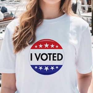 Op de afbeelding: Wit T-shirt met een cirkelvormige afbeelding met de tekst "I VOTED". De afbeelding is rood, wit en blauw met witte sterren. De persoon draagt het shirt voor boten en een houten steiger.