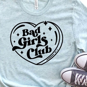 Può includere: Una maglietta blu chiaro con una grafica a forma di cuore nero che dice "Bad Girls Club". La maglietta è abbinata a pantaloncini di jeans e scarpe da ginnastica grigie.