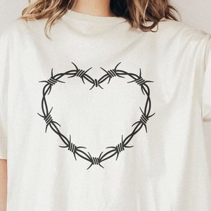 Könnte beinhalten: Ein weißes T-Shirt mit einem schwarzen Stacheldraht-Herz-Design. Das Shirt ist oversized und hat eine lockere Passform.