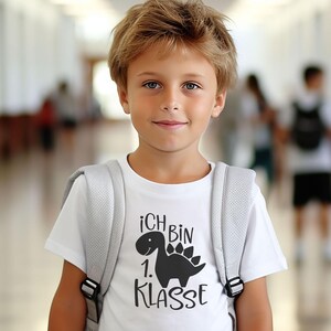 May include: A white short-sleeved t-shirt featuring a black graphic of a dinosaur and the text "ICH BIN 1. KLASSE" printed on the front. The shirt is worn with a light grey backpack with visible shoulder straps and black plastic buckles.