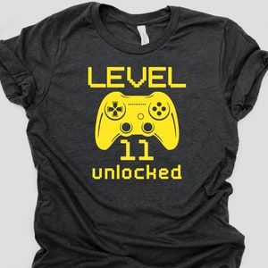 May include: A dark gray t-shirt with a yellow graphic of a video game controller and the text "LEVEL 11 unlocked".