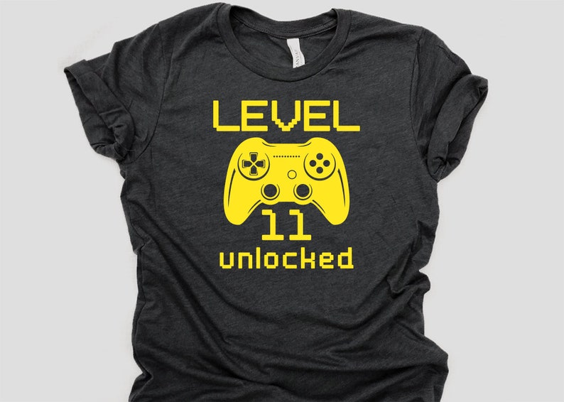 May include: A dark gray t-shirt with a yellow graphic of a video game controller and the text "LEVEL 11 unlocked".