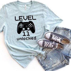 May include: A light blue t-shirt with black text that reads "LEVEL 11 unlocked" and a black video game controller graphic.  The shirt is paired with a pair of dark gray sneakers and blue denim shorts.