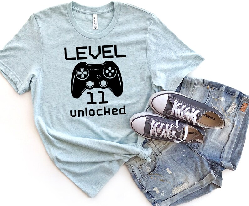 May include: A light blue t-shirt with black text that reads "LEVEL 11 unlocked" and a black video game controller graphic.  The shirt is paired with a pair of dark gray sneakers and blue denim shorts.