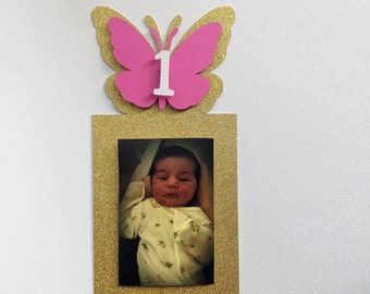 May include: A gold glitter picture frame with a pink glitter butterfly on top. The butterfly has a white number "1" in the center. A photo of a baby is inside the frame.