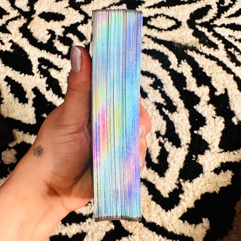 Puede incluir: Una baraja de cartas con un dise&ntilde;o de fondo hologr&aacute;fico iridiscente. Las cartas est&aacute;n apiladas y se sostienen en una mano.
