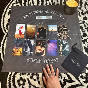 Puede incluir: Un tapiz gris con el texto "I HAVE NO KNOWLEDGE, JUST SENSES." El tapiz presenta un despliegue de cartas de tarot con im&aacute;genes de fuego, una mujer con los ojos vendados, una corona y una persona con los brazos en alto. El tapiz est&aacute; sobre una superficie con estampado blanco y negro. Una bolsa negra con el texto "Rare Rebel" se encuentra en la esquina inferior derecha de la imagen.