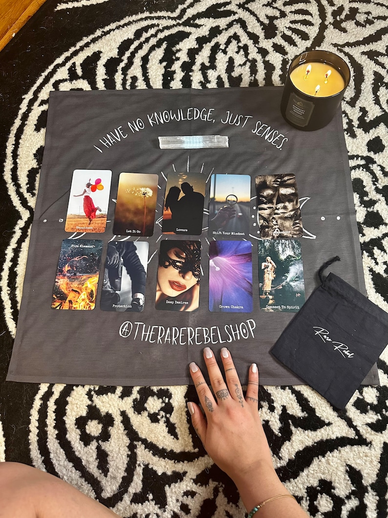Puede incluir: Un tapiz gris con el texto "I HAVE NO KNOWLEDGE, JUST SENSES." El tapiz presenta un despliegue de cartas de tarot con im&aacute;genes de fuego, una mujer con los ojos vendados, una corona y una persona con los brazos en alto. El tapiz est&aacute; sobre una superficie con estampado blanco y negro. Una bolsa negra con el texto "Rare Rebel" se encuentra en la esquina inferior derecha de la imagen.