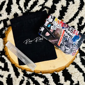 Puede incluir: Una bolsa de tela negra con las palabras "Rare Rebel" impresas en blanco. Una varita de cristal blanco est&aacute; sobre una superficie de madera junto a la bolsa. Una tarjeta con un collage de im&aacute;genes y las palabras "Rare Rebel" est&aacute; sobre la superficie de madera.