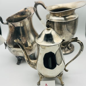 以下が含まれることがあります： 蓋付きの小さなティーポット、クリーマー、ピッチャーなど、アンティークの銀食器のコレクション。 ティーポットには装飾的なディテールがあり、3つの小さな足の上に立っています。 クリーマーとピッチャーにも装飾的なハンドルと注ぎ口が付いています。