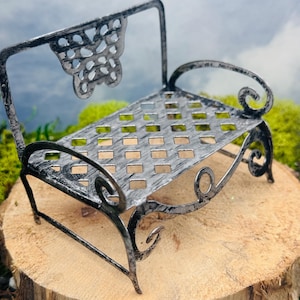 May include: A miniature metal bench with a distressed silver finish. The bench has a decorative back with cut-out details and a rectangular seat. The scrolled armrests and legs add a whimsical touch. The bench is placed on a wooden slice.