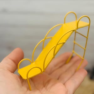 May include: A miniature yellow metal slide with curved railings and a ladder. The slide is held in a hand, showcasing its small size. The bright yellow color contrasts with the background, highlighting the toy's playful design.
