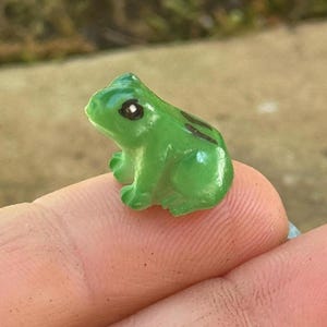 May include: A small, green frog figurine with black markings and large eyes. The frog is perched on a finger, showcasing its miniature size and detailed features. The figurine is a vibrant green color.