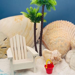 Peut inclure: Scène de plage miniature avec une chaise Adirondack en bois blanc, deux palmiers, un seau rouge avec une poignée jaune et des coquillages sur une surface sablonneuse. Le fond est bleu uni.