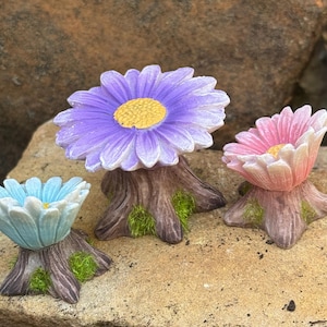 May include: Three miniature flower sculptures in blue, purple, and pink. Each flower sits atop a tree trunk base with green moss accents. The sculptures are arranged on a light brown stone surface.