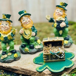 Puede incluir: Una colección de figuras del Día de San Patricio. Tres figuras de duendes con trajes y sombreros verdes, con detalles dorados, están cerca de un cofre del tesoro lleno de monedas de oro y un trébol. Las figuras están sobre una superficie de piedra.