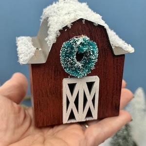 May include: A small wooden red barn with a white door and a green wreath on the front. The roof is covered in white faux snow.