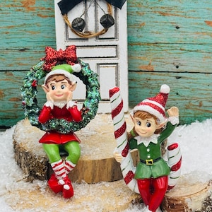 May include: Two Christmas elf figurines. One elf is wearing a red and green outfit and holding a green wreath with a red bow. The other elf is wearing a green and red outfit and holding a candy cane. A miniature white door with a black bow and bells is in the background.