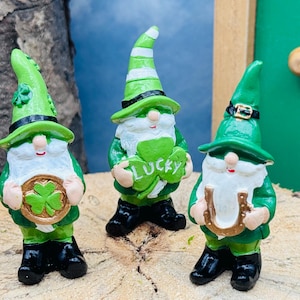 Könnte beinhalten: Drei grüne und weiße Zwergfiguren. Jeder Zwerg trägt einen grünen Hut und eine Jacke, schwarze Stiefel und einen weißen Bart. Einer der Zwerge hält eine Goldmünze mit einem Kleeblatt, ein anderer ein Kleeblatt mit dem Wort "LUCKY" und der dritte ein goldenes Hufeisen.