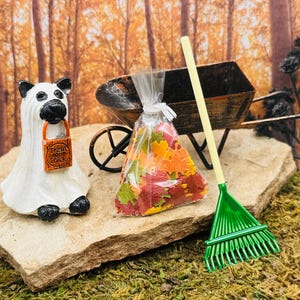 Puede incluir: Una escena en miniatura con temática de Halloween con una figura de fantasma blanco que sostiene una bolsa de truco o trato. Una pequeña carretilla, una bolsa de hojas coloridas y un rastrillo verde completan la exhibición otoñal.