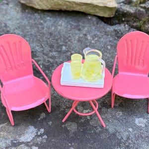 Pode incluir: Um conjunto de pátio em miniatura rosa brilhante. O conjunto inclui duas cadeiras, uma mesa redonda e uma bandeja com um jarro e dois copos. O jarro e os copos parecem estar cheios de um líquido amarelo.