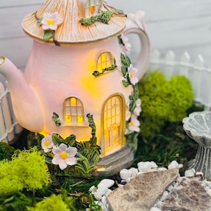 May include: A pink teapot-shaped fairy house with a tan roof, decorated with flowers and greenery. The house features glowing windows and a door, set in a miniature garden with moss, rocks, and a white picket fence. A charming decorative item.