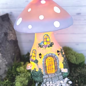 May include: A whimsical mushroom-shaped lamp with a glowing orange cap dotted with white circles. The base is a fairy house with windows, a door, and stone steps, surrounded by greenery. The lamp is designed to illuminate a room.
