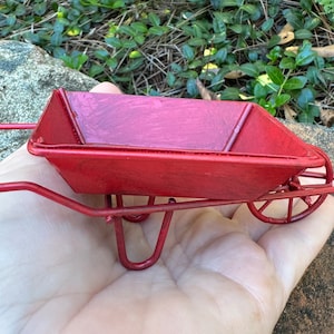 Könnte beinhalten: Ein kleiner roter Metall-Gärtnerwagen mit einem Griff. Der Wagen ist etwa 15 cm lang und 10 cm breit.